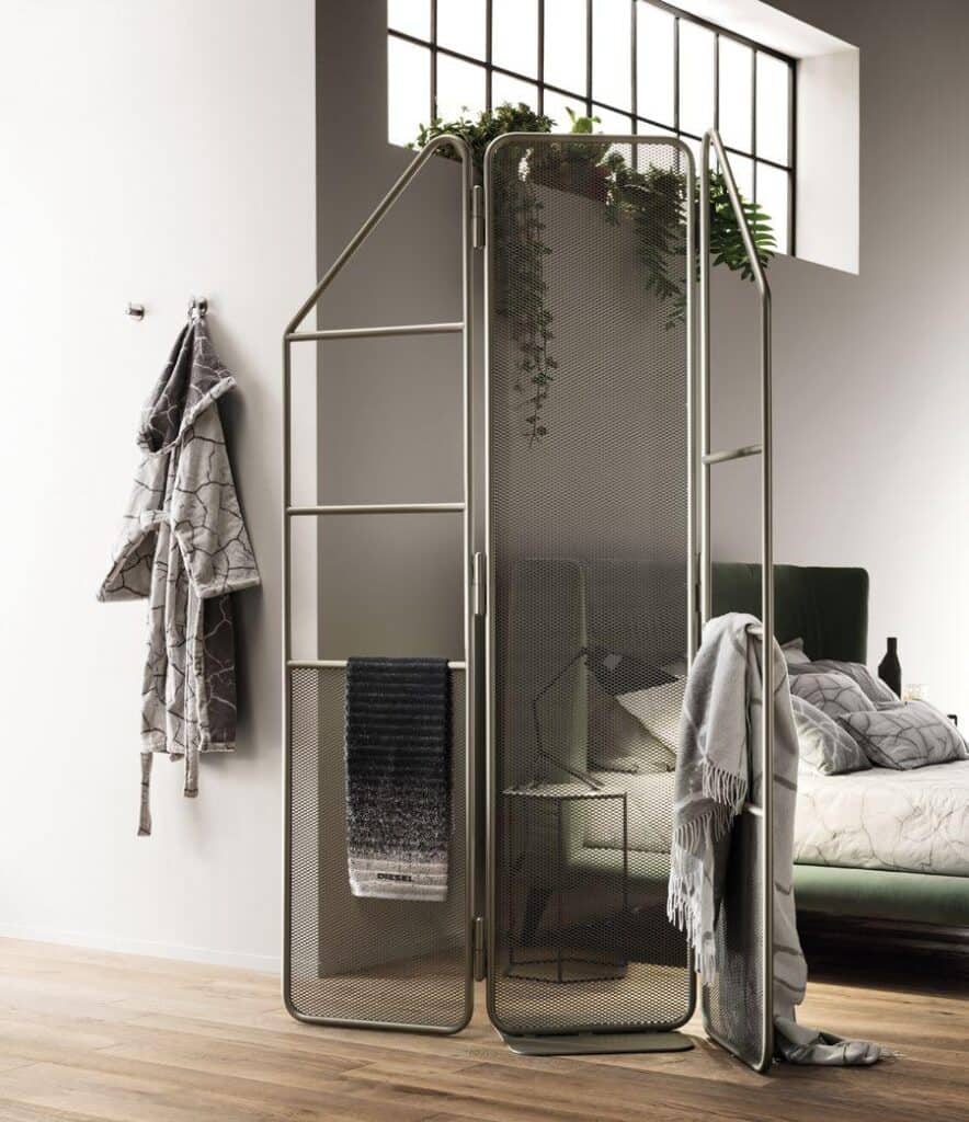 A modern loft-style bedroom with a metal room divider featuring mesh panels. The divider has towels draped over it, with a bed in the background and plants placed atop the divider under a large window. A patterned robe hangs on the wall.