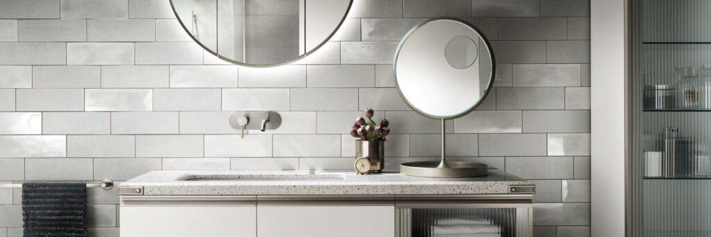 Modern bathroom with a terrazzo countertop, circular mirror above, a freestanding magnifying mirror, metal faucet, and decorative tiles.