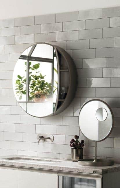 A bathroom with a round wall mirror and a smaller round tabletop mirror, featuring a reflection of potted plants, against a tiled wall.