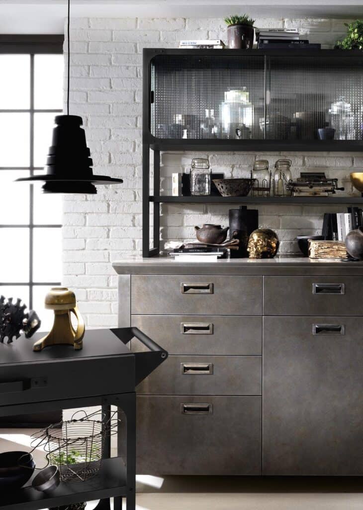 Industrial-style kitchen with metal cabinets, open shelves displaying jars and decor, black pendant light, and wheeled cart against a white brick wall.