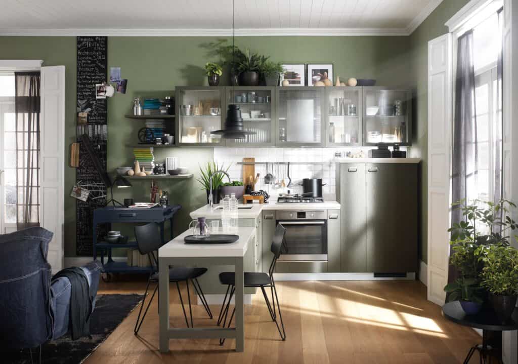 Modern kitchen with green walls, black shelving, glass-front cabinets, a white island, and various plants.
