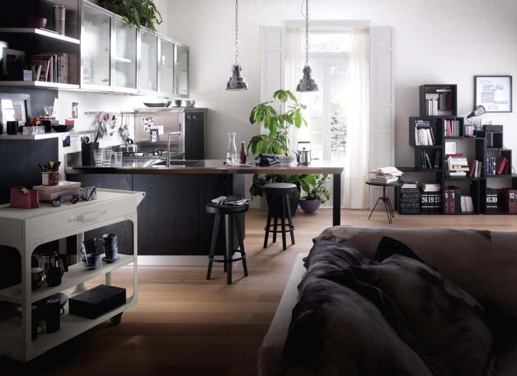 Modern open-plan living and kitchen space with black cabinetry, shelving, island with stools, a rolling cart, sofa, and large plant near a window.