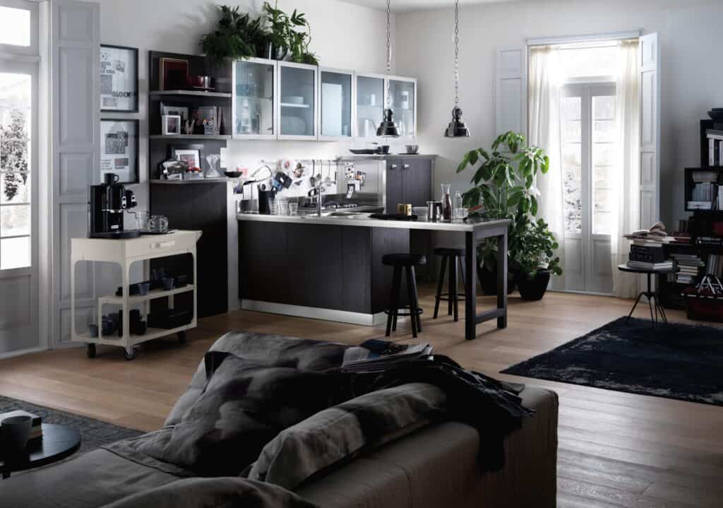 Stylish modern kitchen with dark cabinets, bar stools, a coffee cart, and plants near large windows with sheer curtains letting in natural light.