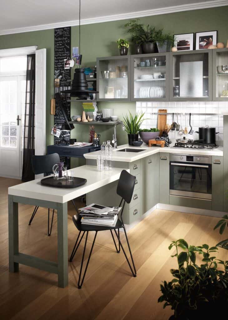 Modern kitchen with green cabinets, wooden floor, black chairs, and a white countertop. There&#039;s a built-in oven, gas stove, and a chalkboard wall. Plants and books are displayed on shelves.