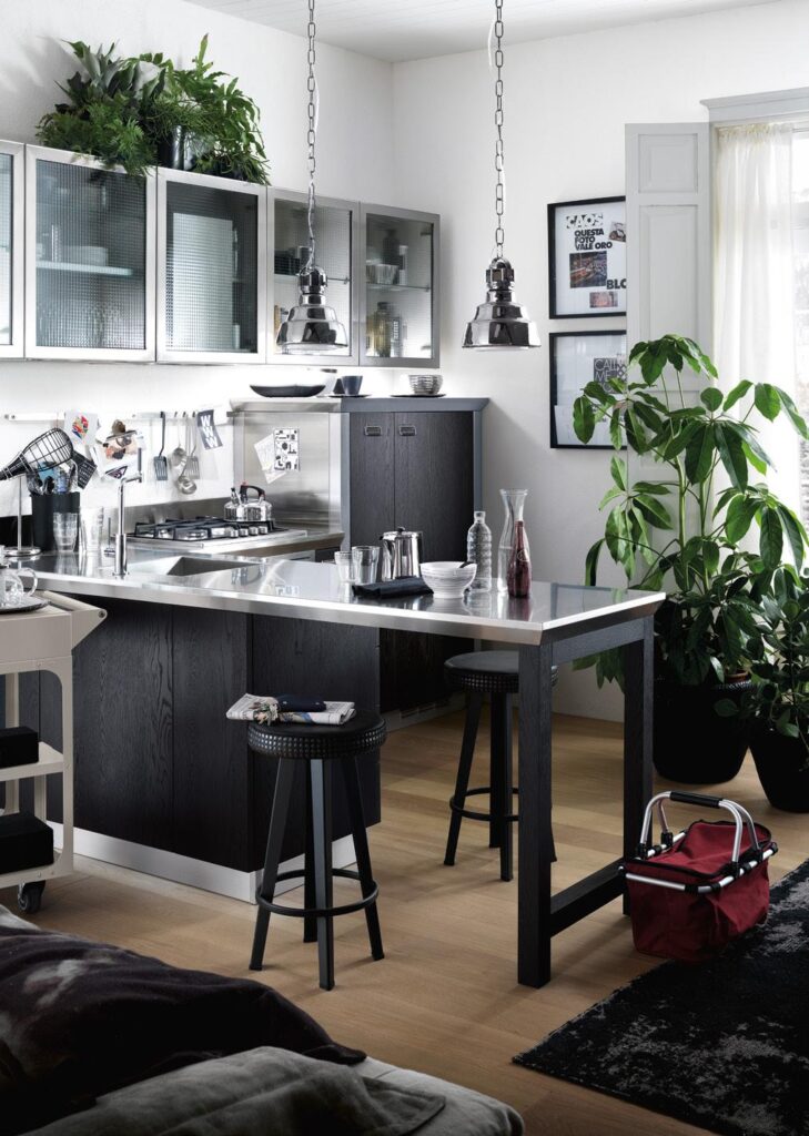 A modern kitchen with dark cabinets, overhead glass cabinets, and stainless steel fixtures. A central island has two black stools, pendant lights above, and decorative plants placed nearby.