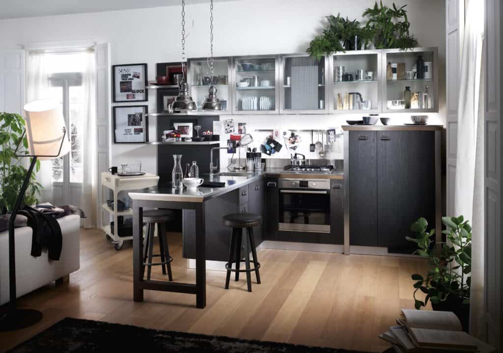 Modern kitchen interior with dark wood cabinets, stainless steel appliances, and hanging lights. There are stools at a breakfast bar and plants adding greenery to the space.