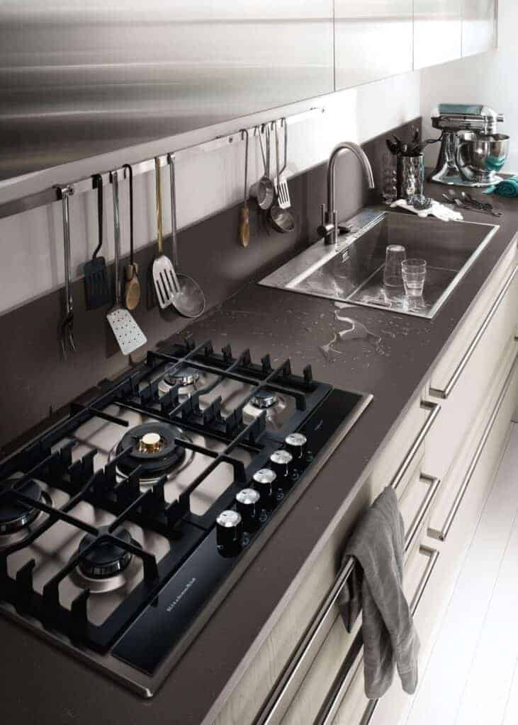 Modern kitchen with a gas stove, utensils hanging above, a sink with water droplets on the counter, and a mixer in the background.