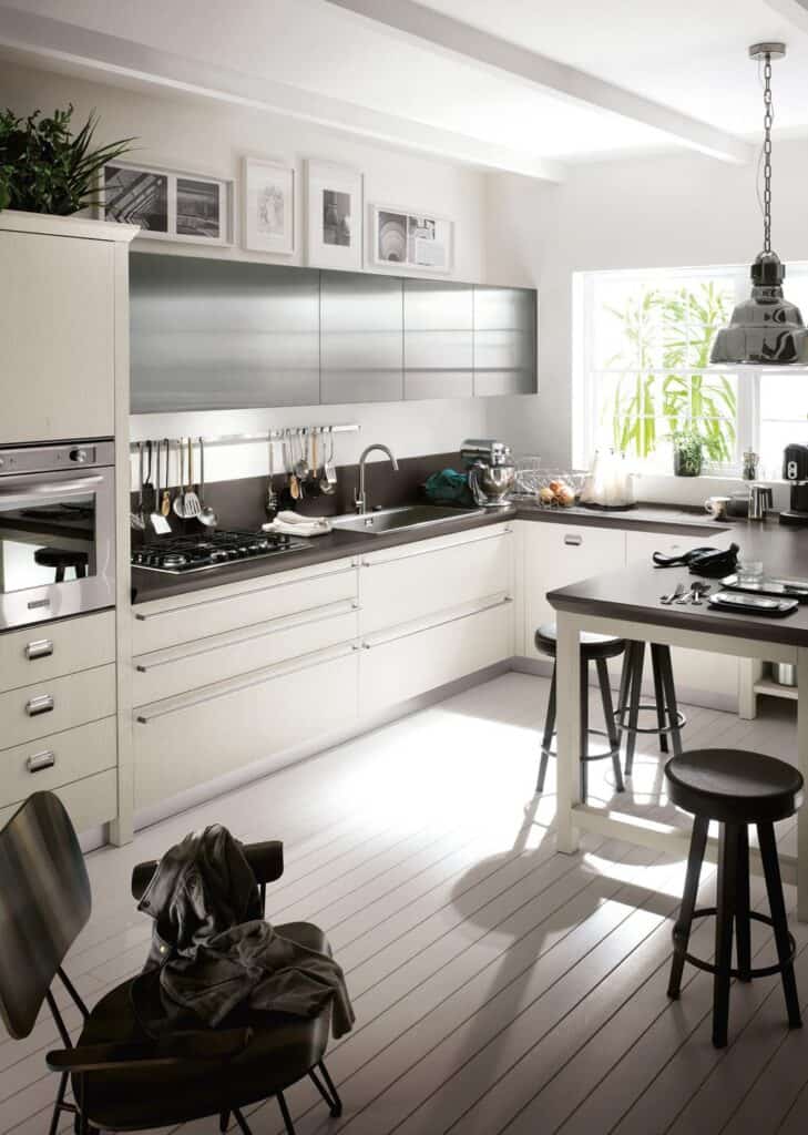 Modern kitchen with white cabinetry, a black countertop, and stainless-steel appliances. A small island with barstools is in the center, and framed pictures decorate the upper wall. Natural light streams in through a large window.