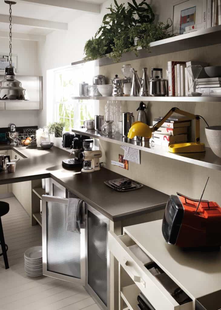 Modern kitchen with open shelves displaying various kitchenware, books, and plants; a countertop with coffee makers, a red TV, and a yellow lamp; frosted glass cabinet doors below.
