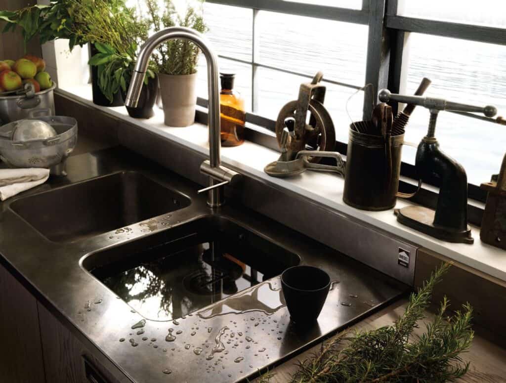 Kitchen sink with water droplets, surrounded by potted plants, antique utensils, and a window view.