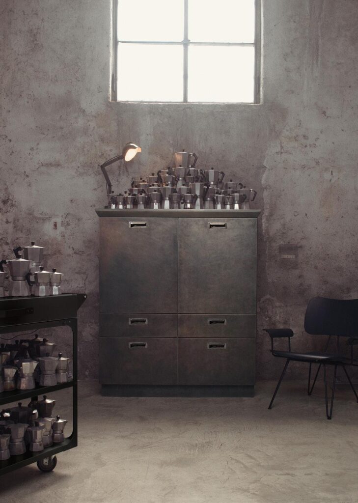 A metal cabinet topped with a large collection of moka pots, set against a rustic concrete wall with a window above and a chair on the side.