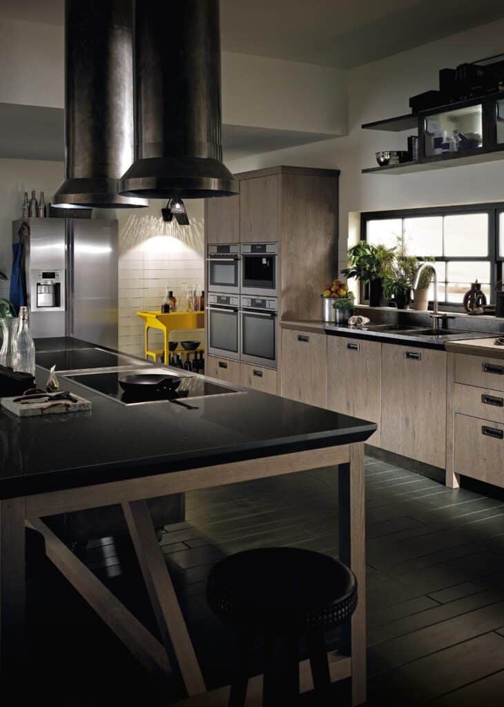 Modern kitchen with wooden cabinets, black countertops, stainless steel appliances, a central island with stools, and hanging industrial-style vents.