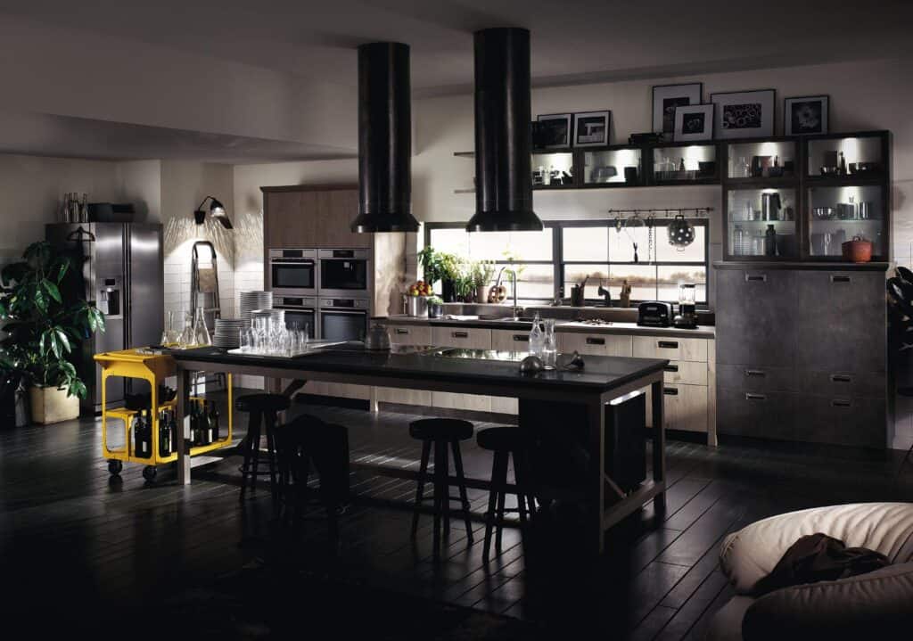 Modern industrial kitchen with dark wood floors, a long central island with stools, open shelving holding various kitchen items, two large ceiling vents, a stainless steel refrigerator, and a yellow cart holding bottles.