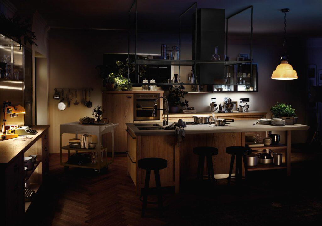 A dimly lit modern kitchen with a central island, stools, hanging light fixture, and various kitchen utensils and plants on shelves.