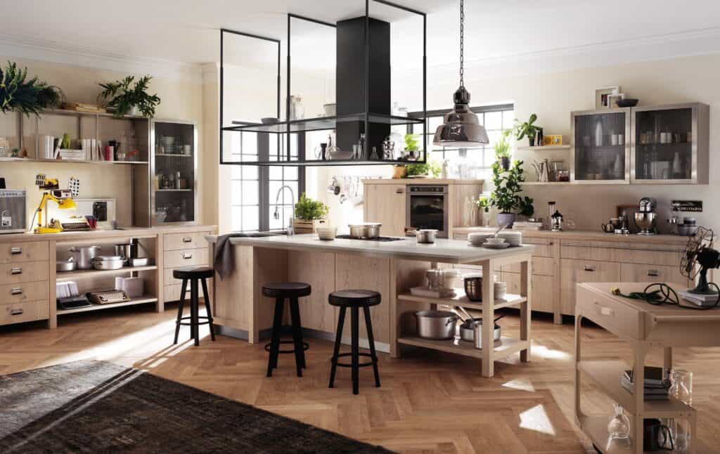 Modern kitchen interior with light wood cabinets, a central island with stools, open shelving, and various kitchen appliances and plants.