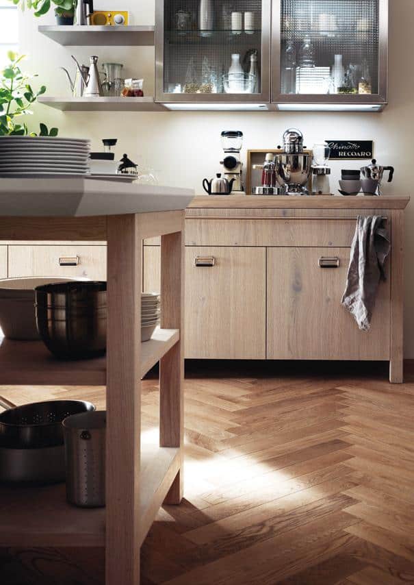 Modern kitchen with wooden cabinets, open shelves displaying kitchenware, and a herringbone wood floor.