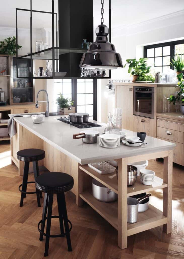 Modern kitchen interior with a central island, black pendant light, wooden cabinetry, stools, and open shelves displaying white dinnerware and metal cookware.