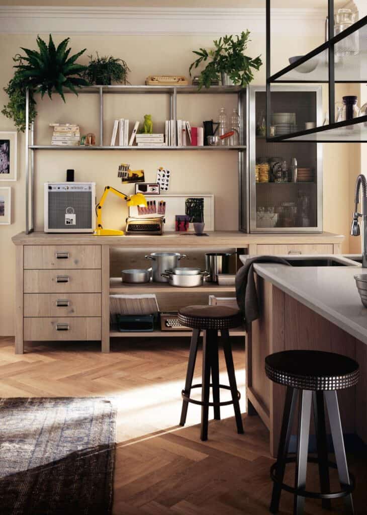 Cozy kitchen corner with wooden shelves displaying books, plants, and decor; a counter with a typewriter and a yellow lamp; metal stools by an island; and a cabinet with pots and kitchenware.