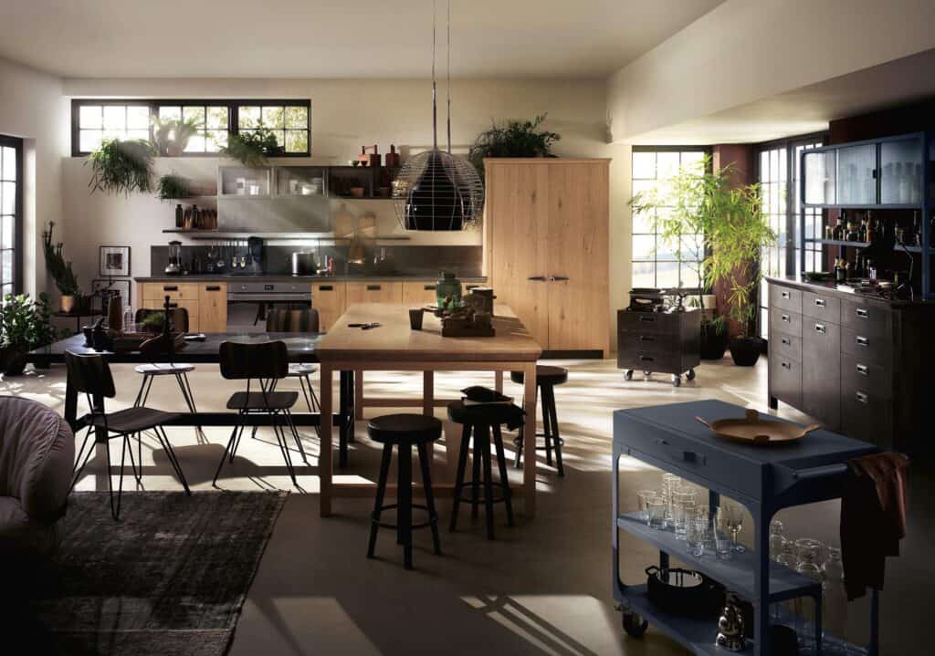 Modern kitchen with wooden cabinets, a large island, and black chairs. Indoor plants and ample natural light brighten the space.