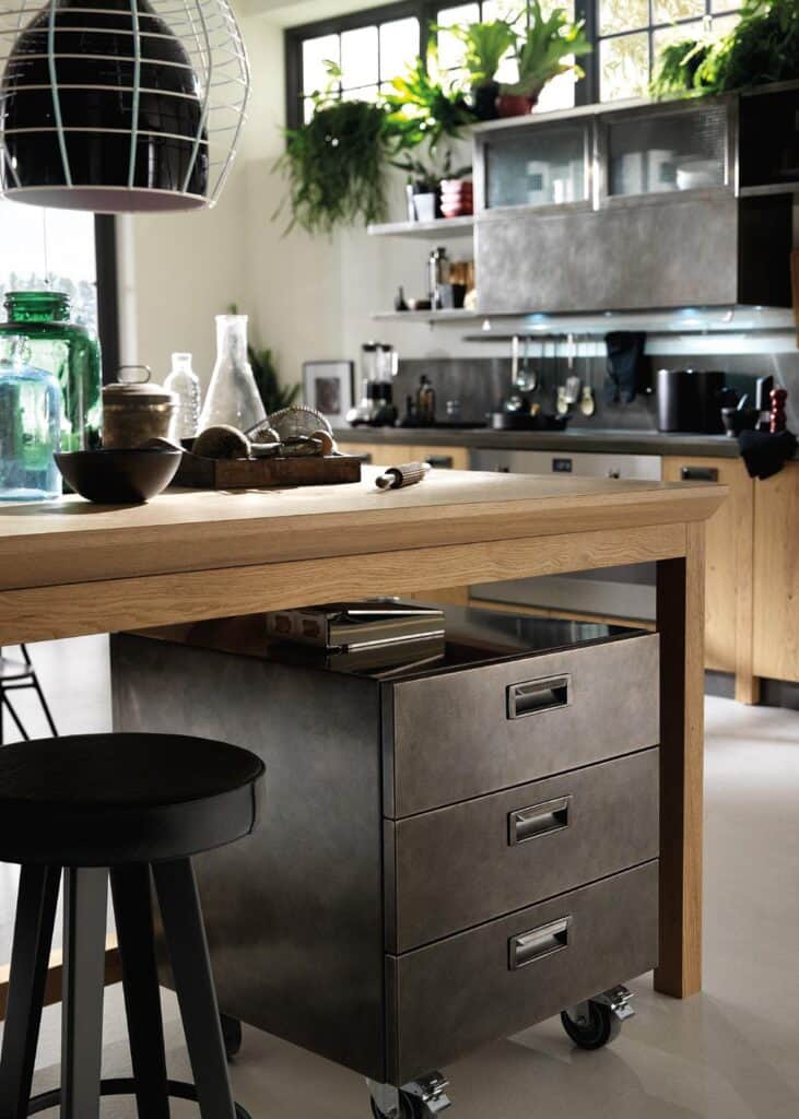 Modern kitchen interior with a wooden island, metal drawer unit on wheels, overhead lights, and plants on shelves.