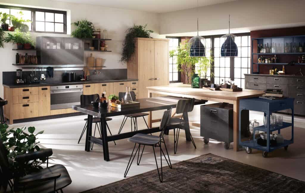 Modern kitchen with wooden cabinets, open shelves, plants, and black dining table with chairs. Large windows provide natural light, and pendant lights hang above a wooden island.