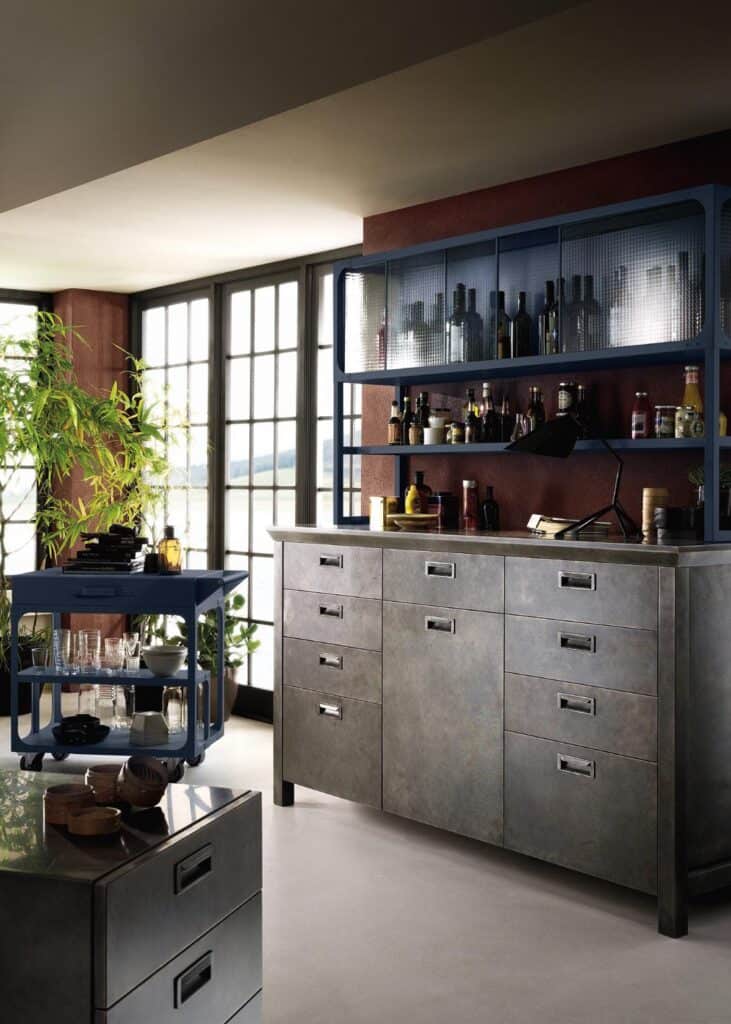 Modern kitchen interior with large windows, metallic cabinets, a bar cart with glassware, and a potted plant.