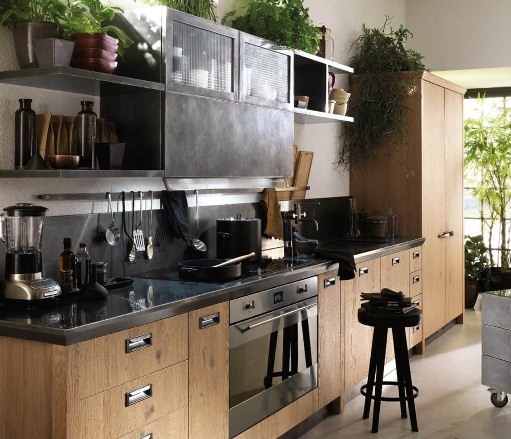 Modern kitchen with wooden cabinets, black countertops, stainless steel appliances, hanging utensils, potted plants, and a stool.
