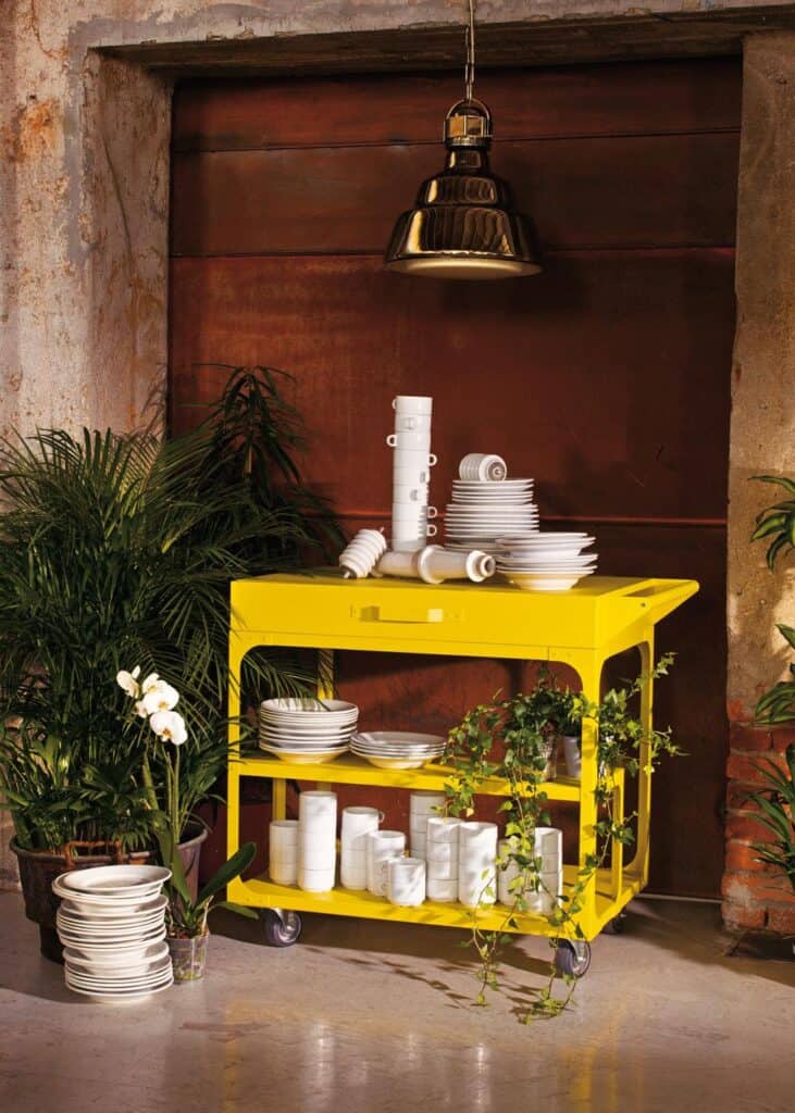 A yellow rolling cart stacked with white dishes and mugs, surrounded by green plants and flowers, under a metallic pendant lamp in an industrial setting.
