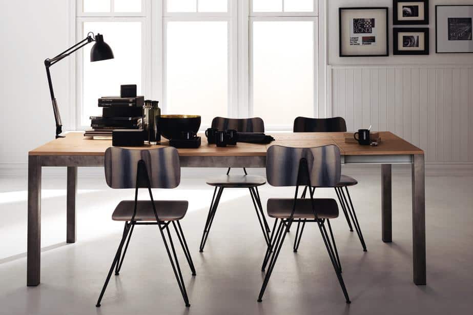 A modern dining room with a wooden table, six chairs, a desk lamp, books, bowls, and mugs, set against large windows and framed photos on the wall.