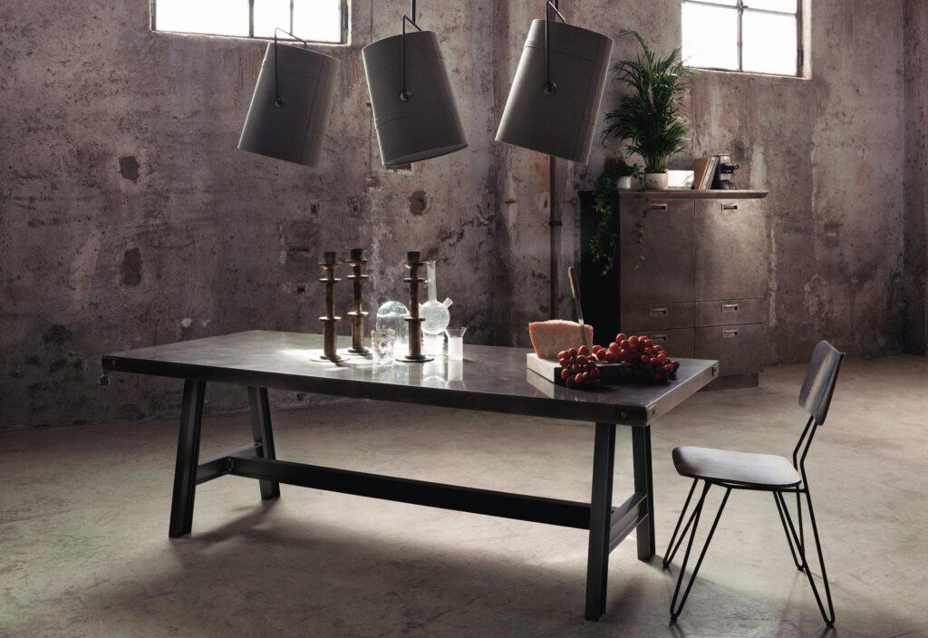 Industrial-style room with metal table and chair, concrete walls, pendant lights, and a cabinet; table has candlesticks, glassware, cheese, and grapes.