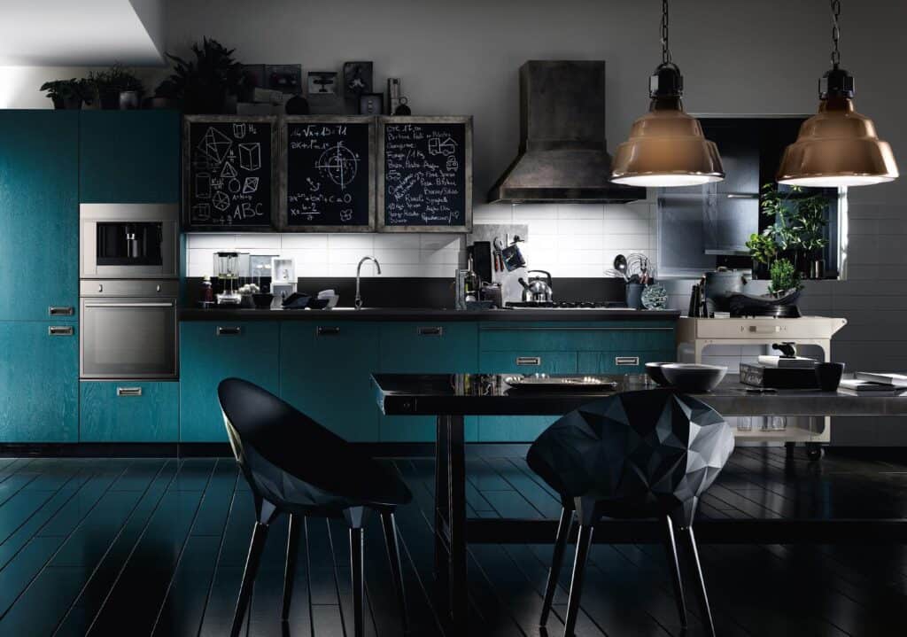 Modern kitchen with teal cabinets, a chalkboard backsplash, industrial pendant lights, black chairs, and a dining table.
