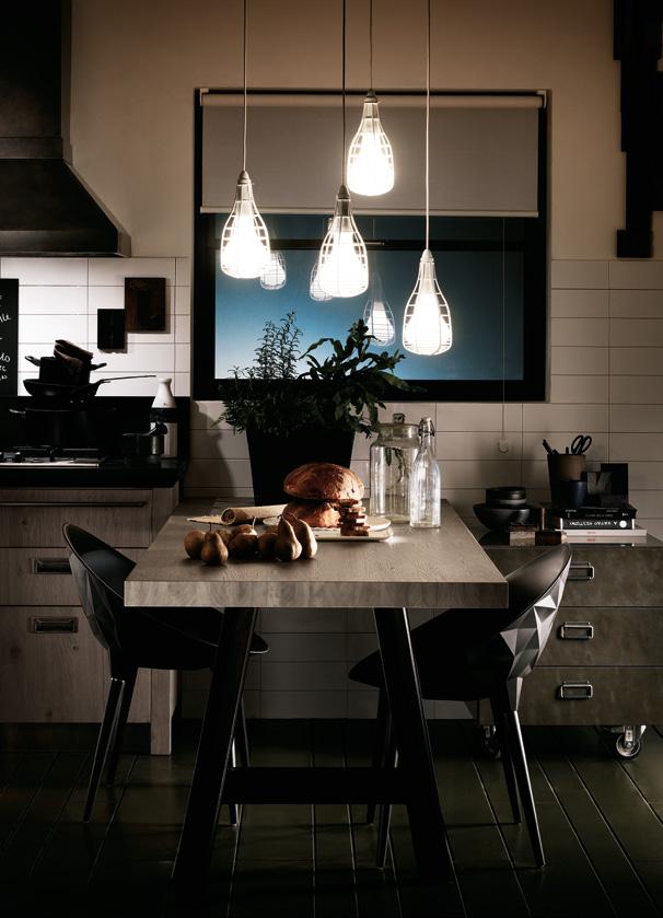Modern kitchen with hanging pendant lights, a wooden table set with bread, potatoes, and glass bottles, black chairs, and a plant near the window.