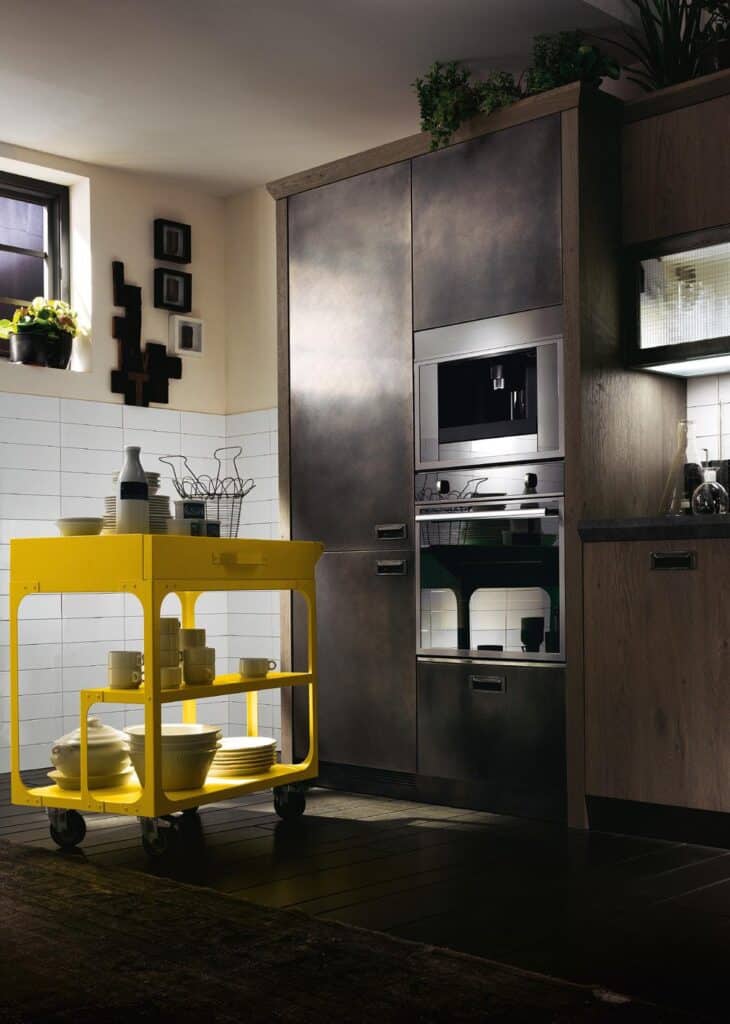 A modern kitchen with metal cabinetry, a built-in oven, and a bright yellow utility cart holding dishes.