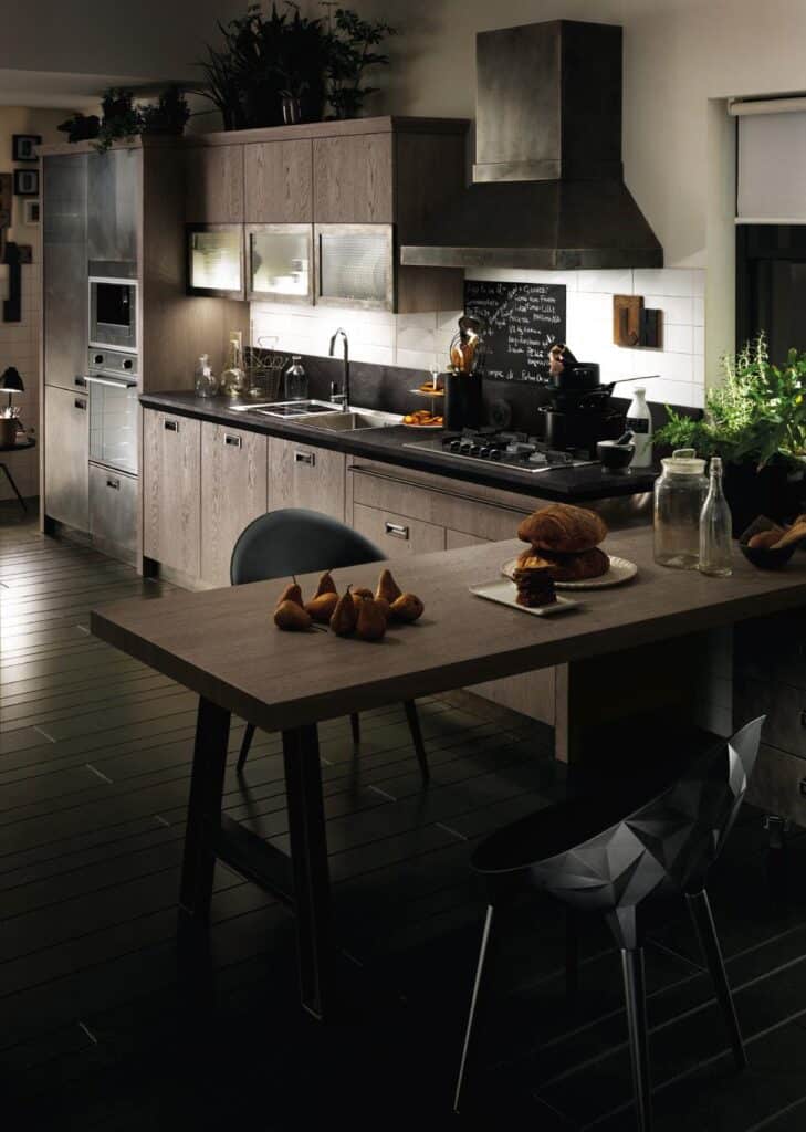 A modern kitchen with wooden cabinets, stainless steel appliances, a dark countertop, and plants. A table in the foreground has bread, figs, and glass bottles.