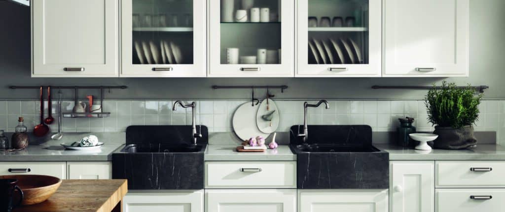 Modern kitchen with black double sinks, white cabinets, glass-fronted upper cabinets displaying dishes, herbs on the counter, and hanging utensils.