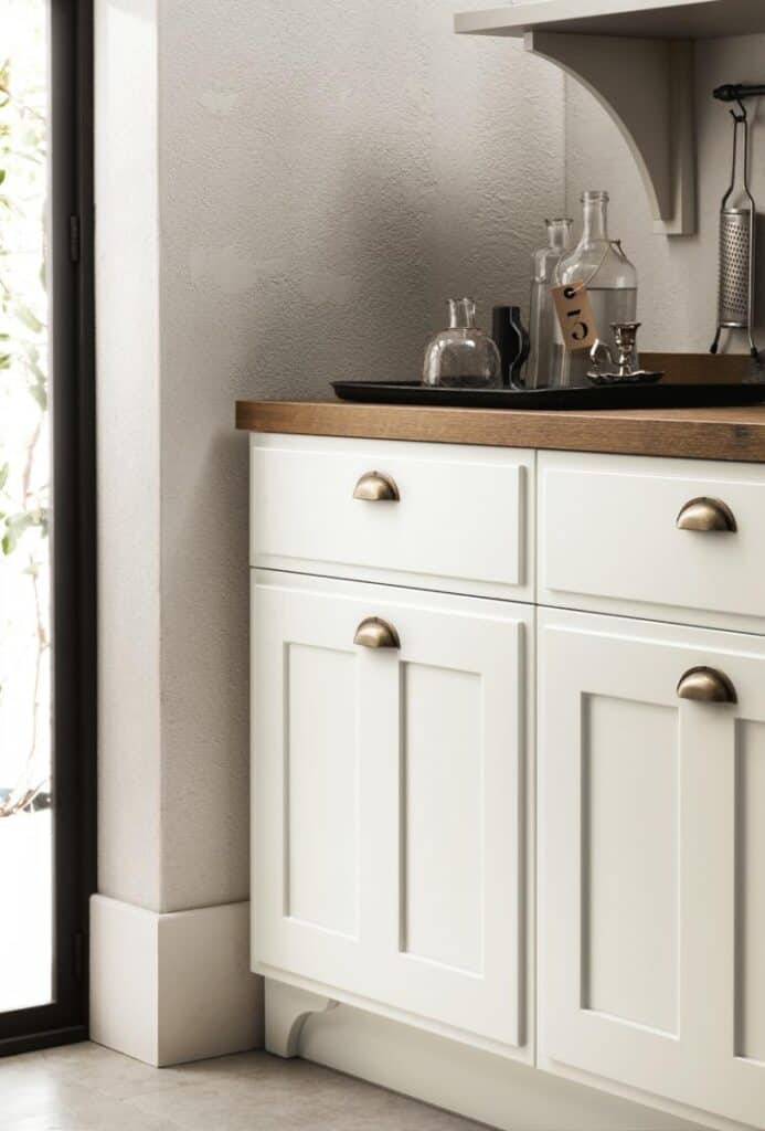 White kitchen cabinets with brass handles, a wooden countertop, and glass bottles on top, next to a window.