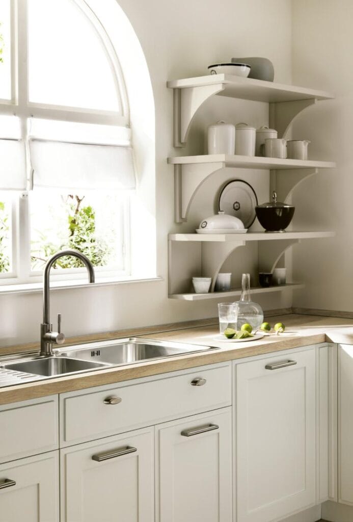 A bright kitchen with white cabinetry, a stainless steel sink, open shelving with kitchenware, and a window with a view of greenery.