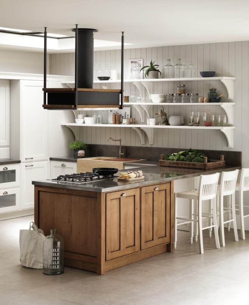 Modern kitchen with a wooden island featuring a stove and sink, white chairs, open shelving with glassware and plants, and light gray walls.