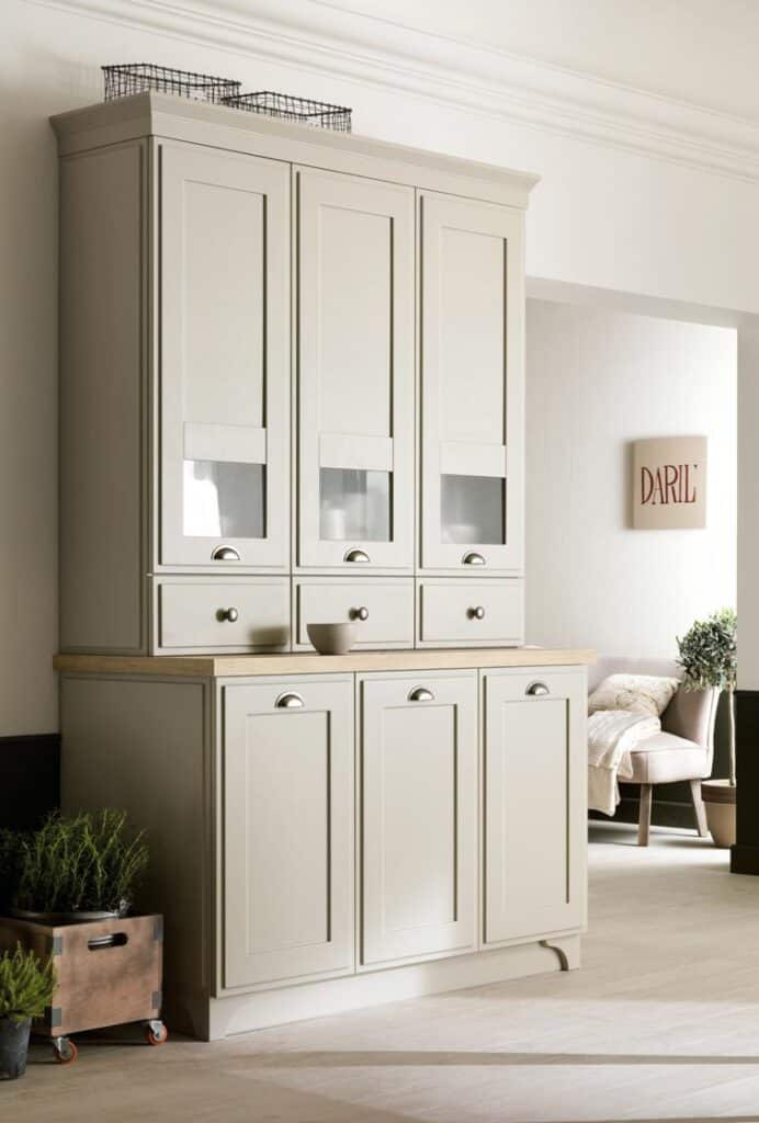 Cream-colored kitchen cabinet with upper glass panels and silver handles, wooden countertop, plants in a wooden container, and a chair in the background.