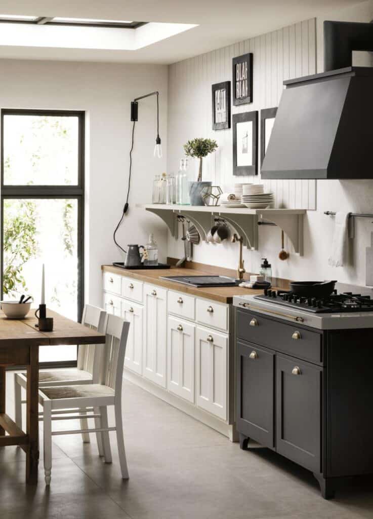 Modern kitchen with white cabinets, wooden countertop, black stove, and open shelving. A wooden dining table with white chairs is on the left. The room has a large window and wall decor.