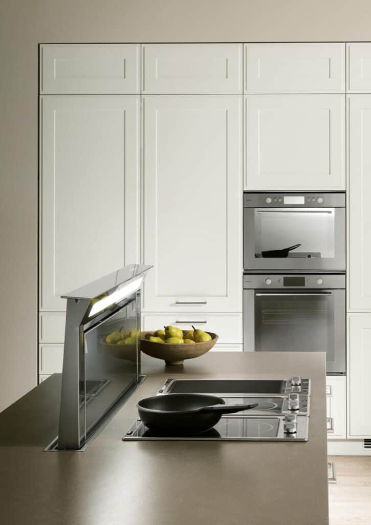 Modern kitchen with induction cooktop, raised vent hood, black frying pan, and bowl of pears. Built-in double oven and white cabinetry in the background.