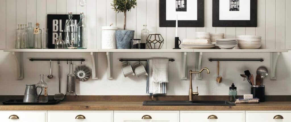 Kitchen countertop with white shelves above, displaying glass bottles, kitchen tools, and stacked dishes; wooden countertop with sink and gold faucet below.