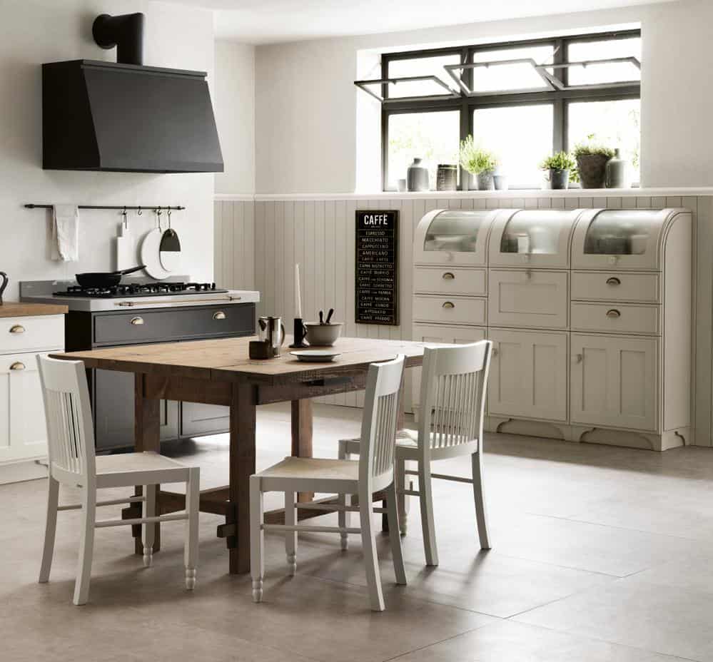 Modern kitchen with a wooden dining table, white chairs, black stove, range hood, and cabinets; large window with potted plants.