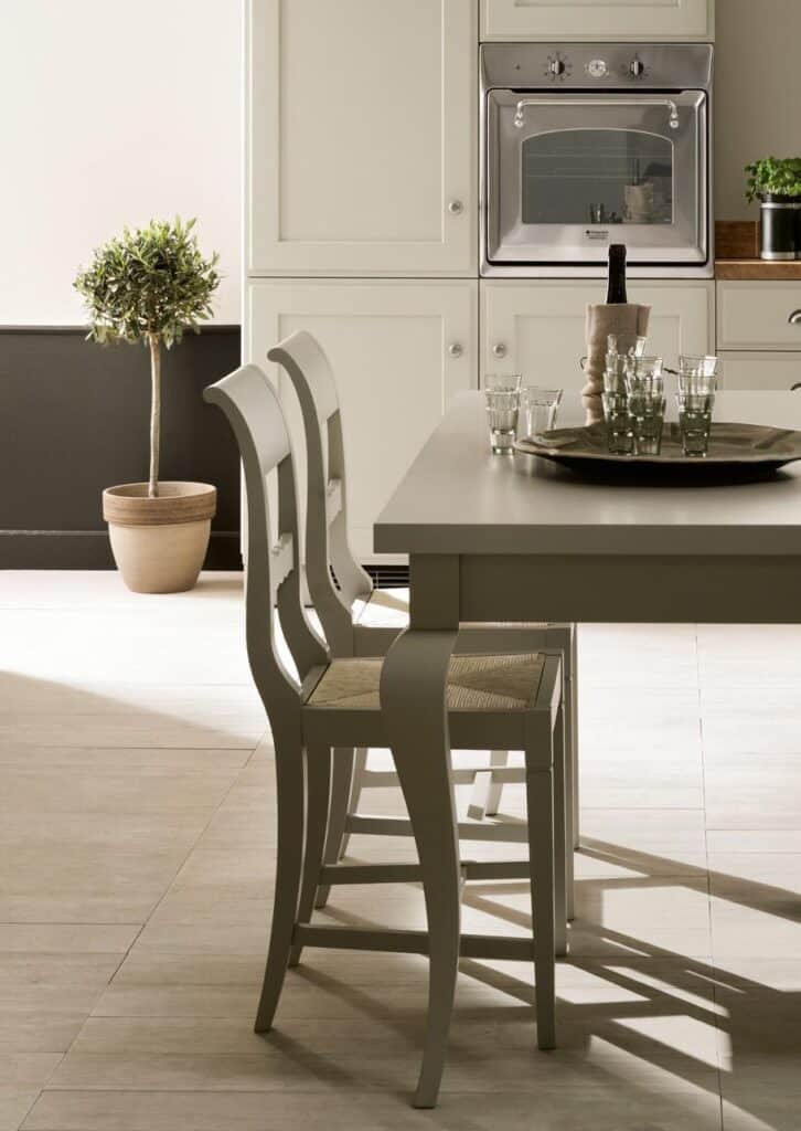 Modern kitchen with white cabinetry, an oven, a dining table with two chairs, a tray with glasses and a wine bottle, and a potted plant.