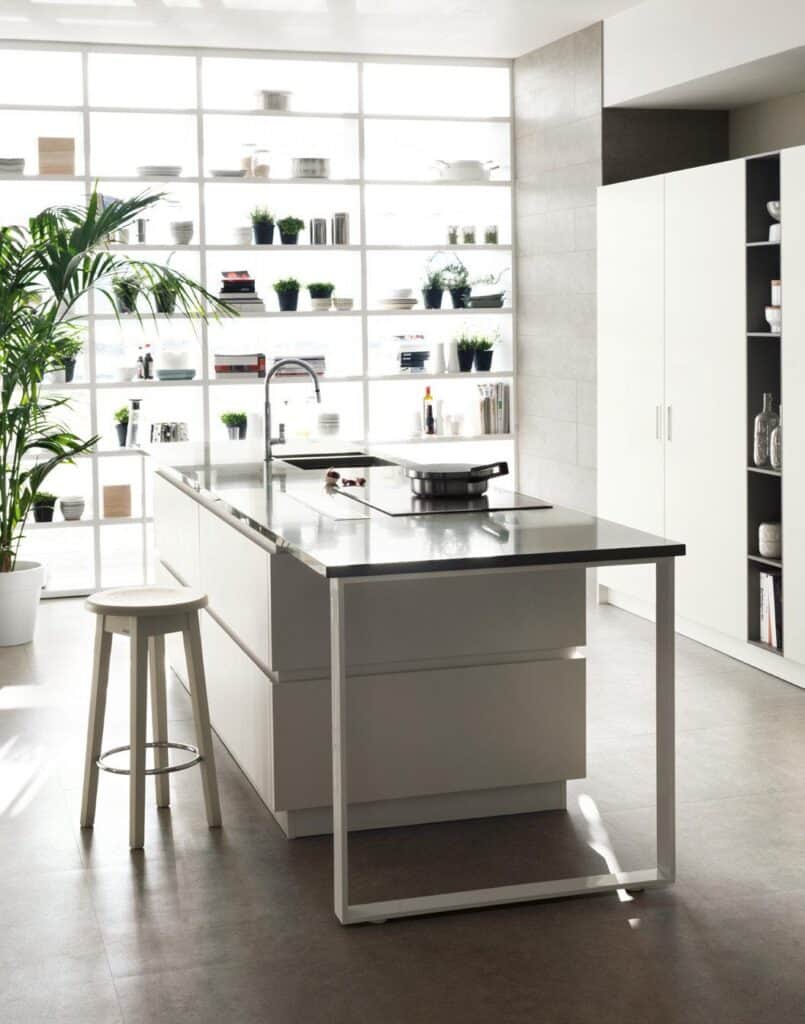 Modern kitchen with a large island, bar stool, and floor-to-ceiling shelving filled with plants and kitchenware.