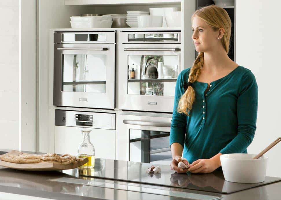 A woman with a braid stands in a modern kitchen, holding garlic near a countertop with a bowl and olive oil bottle. There are stainless steel ovens in the background.