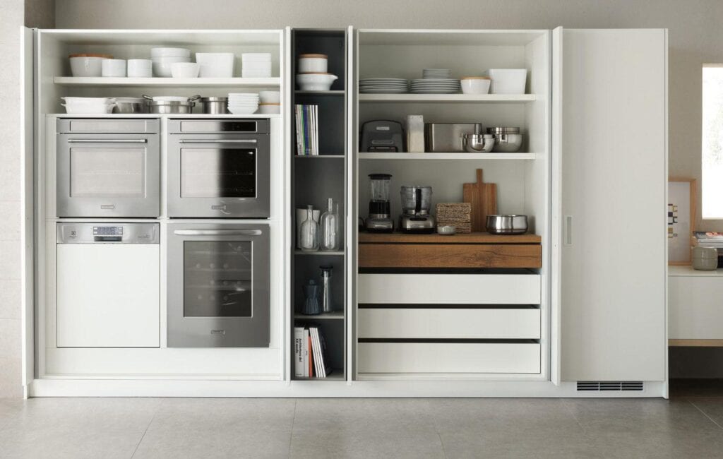 Modern kitchen cabinetry with built-in stainless steel ovens, dishwasher, and open shelves displaying dishes and kitchen appliances.