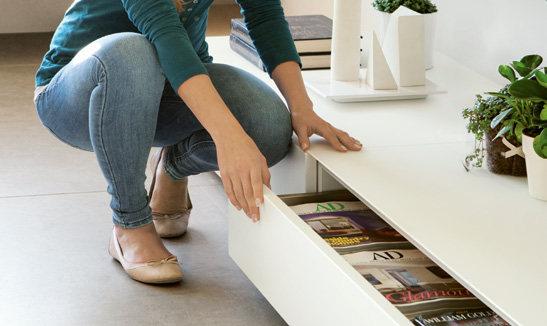 A person in jeans and a teal top opens a white drawer containing magazines, next to a plant on a low shelf.