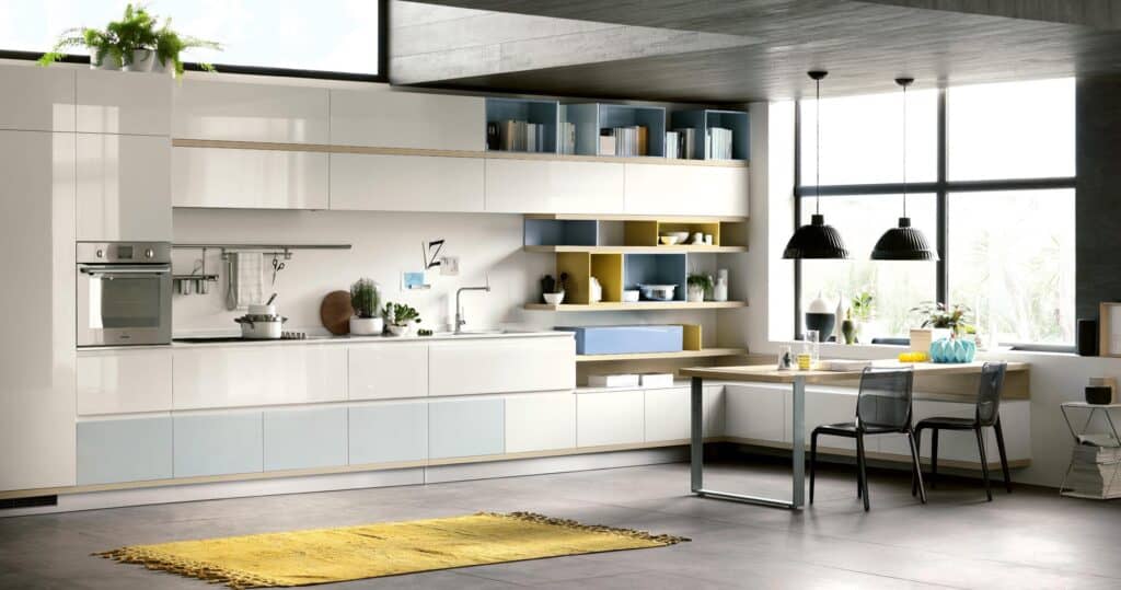 Modern kitchen with sleek white cabinets, open shelving, and a built-in oven. A yellow rug lies on the floor, and a large window lets in natural light. Black pendant lights hang above a dining area with two chairs.