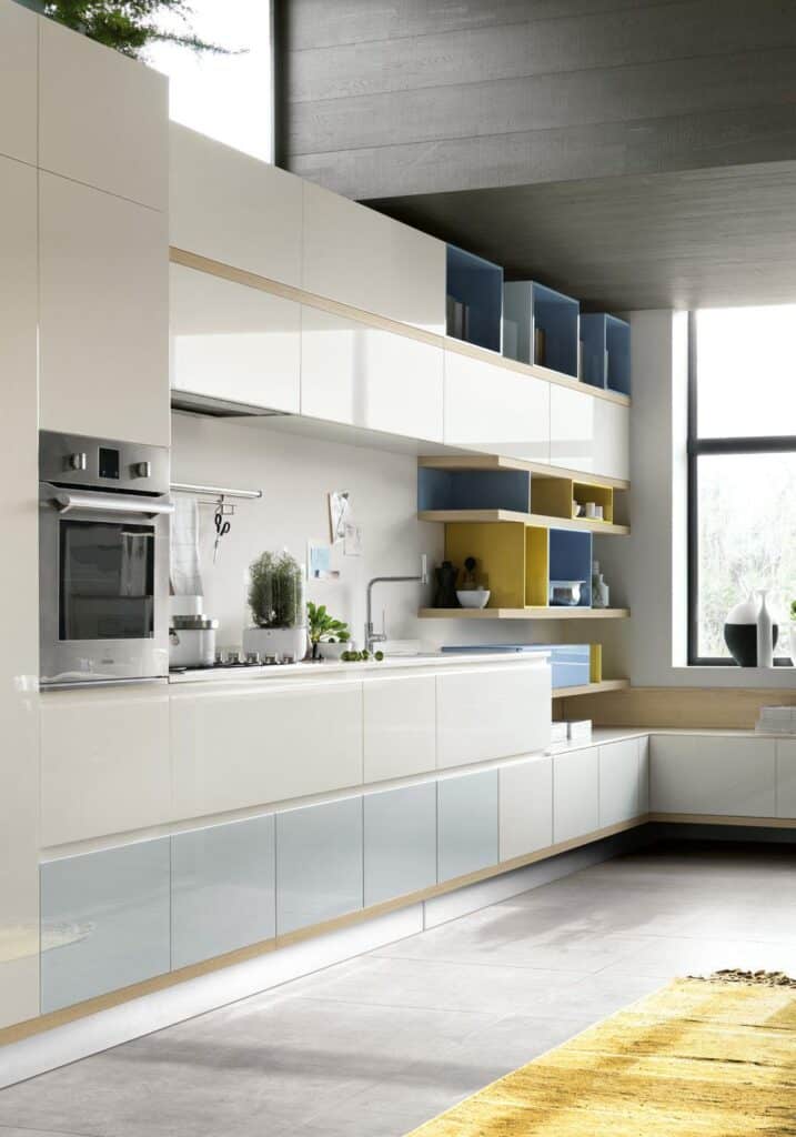 Modern kitchen with sleek white cabinetry, open shelves with blue and yellow accents, stainless steel appliances, and a large window allowing natural light.